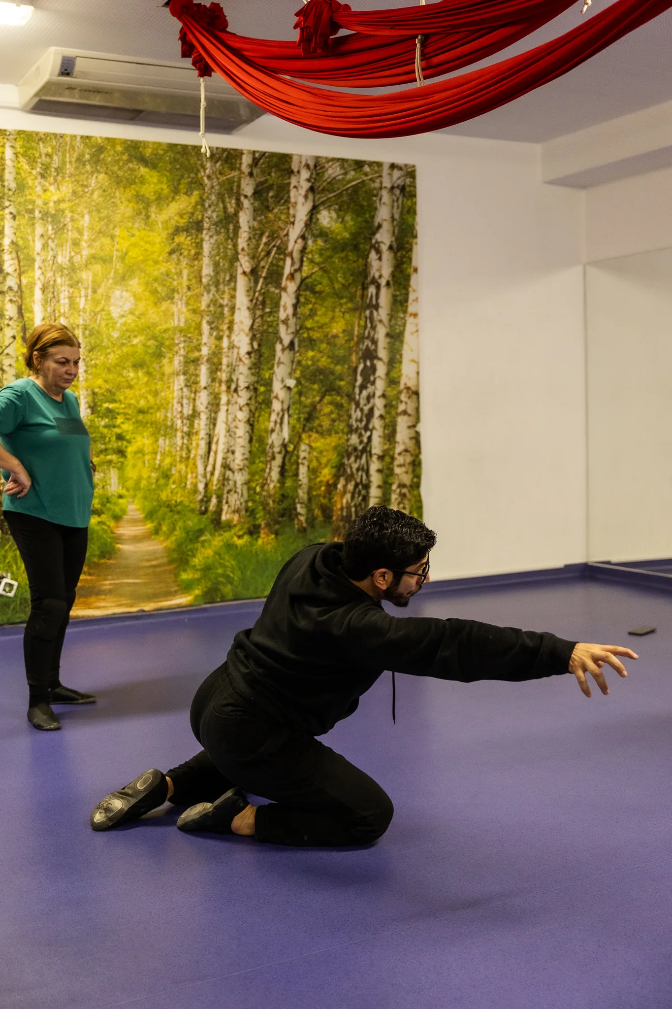 Bailarina ajoelhada, de braços abertos, no estúdio com a parede de floresta